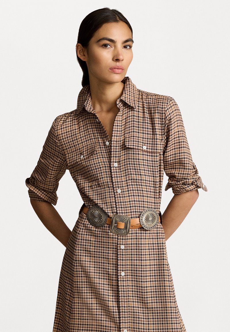 Τα καρό καφέ φόρεμα με ρολαρισμένα μανίκια διαθέτει δύο τσέπες και μια δερμάτινη ζώνη με διακοσμητικές αγκράφες σε ασημί τόνο.