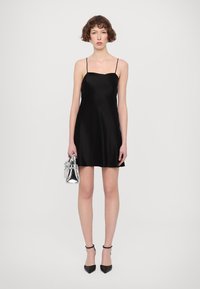 Abito slip in satin nero con spalline sottili, forma a A, texture liscia, accompagnato da una piccola borsa argentata e tacchi neri.