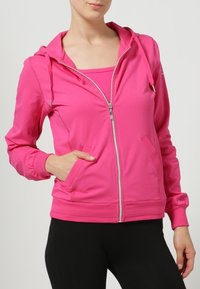 Vaaleanpunainen vetoketjullinen huppari, joka on valmistettu sileästä ja joustavasta kankaasta. Siinä on huppu, kaksi etutaskua ja resorit hihansuissa. Päälle puettu vastaavan värinen toppi.