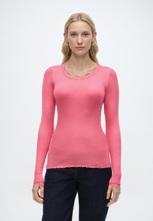 Frau mit langen blonden Haaren, die ein eng anliegendes, rosa langärmeliges Rippenoberteil mit Spitzenbesatz und dunkle Jeans trägt, steht vor einem schlichten weißen Hintergrund.