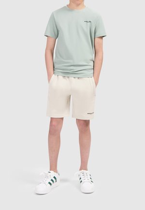 Lichtgroene katoenen t-shirt met zwart logo, gecombineerd met beige katoenen shorts met zakken. Gedragen met witte sneakers met groene strepen.