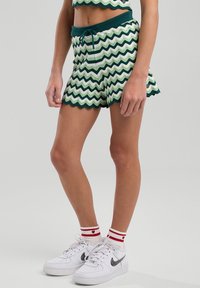 Vihreät ja valkoiset sahalaitakuosiset shortsit, joissa on hapsureunat, naru vyötäröllä, yhdistettynä valkoisiin tossuihin ja punaraitaisiin sukkiin.