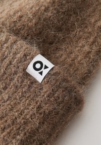 Braunes, grob gestricktes Accessoire aus weichem, strukturiertem Material. Verfügt über ein weißes Etikett mit schwarzem Logodesign. Die Nahaufnahme hebt die Fasern hervor.