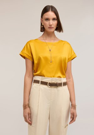 Frau trägt eine gelbe Satin-Kurzarmbluse, beige hoch taillierte Hose mit braunem Gürtel, goldenen Schmuck und einen geraden Bob-Haarschnitt.