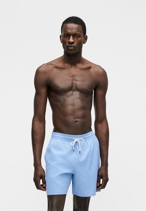 Atletikus férfi, világoskék húzózsinóros fürdőnadrágban áll, egyszerű szürke háttér előtt.