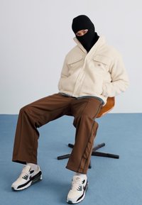 Fleece jakke i creme med to brystlommer, brune træningsbukser med sidelommer og hvide sneakers med sorte detaljer, siddende på en stol.