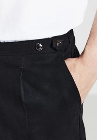 Pantalons en coton noirs avec une ceinture à deux boutons noirs, une poche latérale et une texture en côtes verticales.