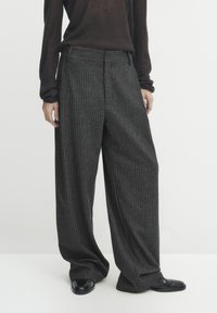 Person wearing loose-fitting dark gray pinstripe trousers with black shoes and a dark long-sleeve top, standing against a plain background.