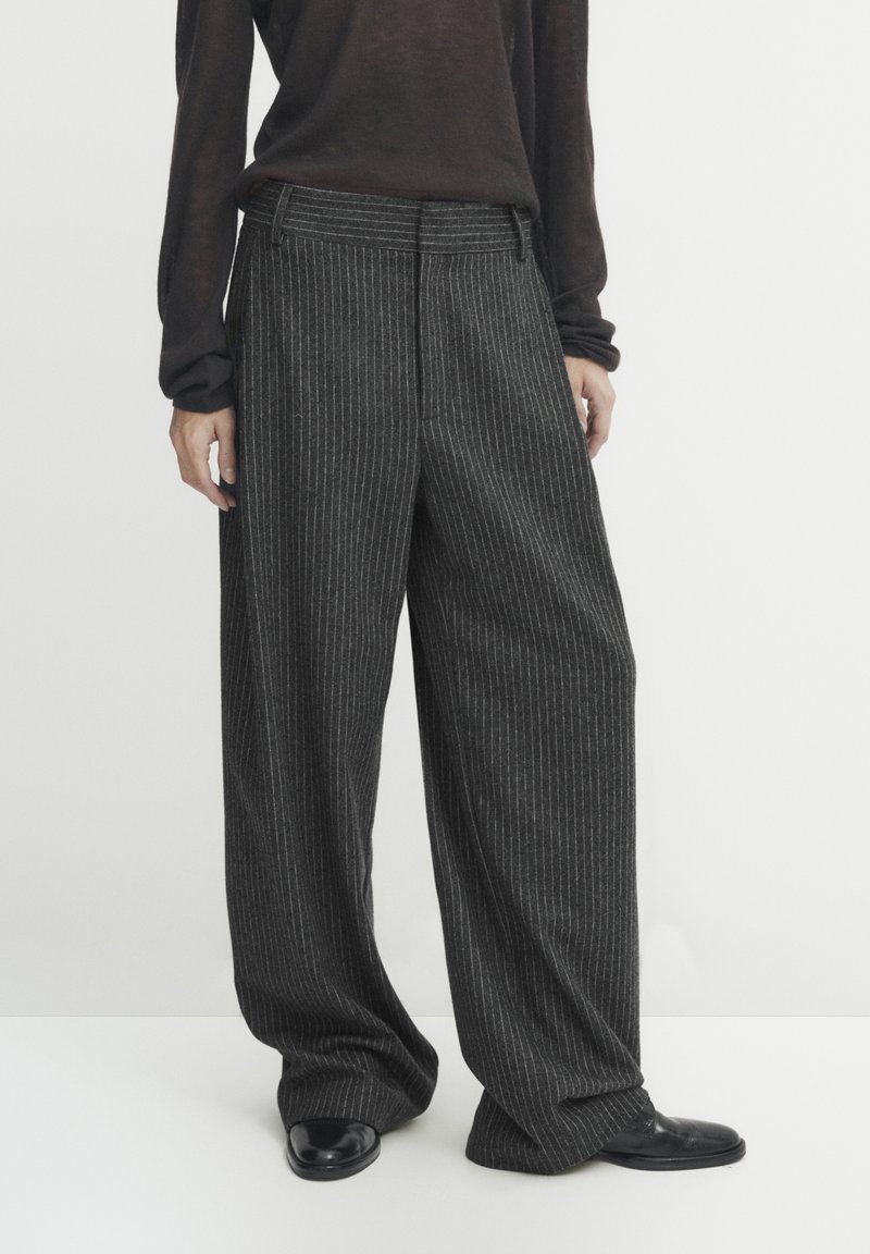 Person wearing loose-fitting dark gray pinstripe trousers with black shoes and a dark long-sleeve top, standing against a plain background.