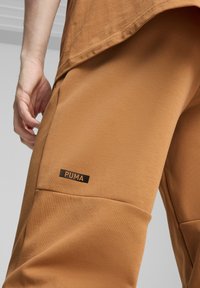 Hnedé športové nohavice vyrobené z mäkkej látky majú výraznú visačku s logom "PUMA". Dizajn obsahuje vrecká a voľný strih.