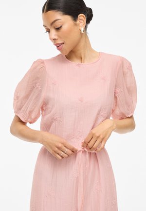 Vaaleanpunainen mekko, jossa on lyhyet puffhihojat, brodeerattuja kukkaketjuja ja vartalonmyötäinen vyötärö, joka on sidottu kangasvyöllä. Pehmeä, teksturoitu kangas.