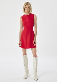 Rotes ärmelloses Kleid mit hohem Ausschnitt und figurbetonter Silhouette, kombiniert mit weißen knielangen Stiefeln und einer glatten Textur.