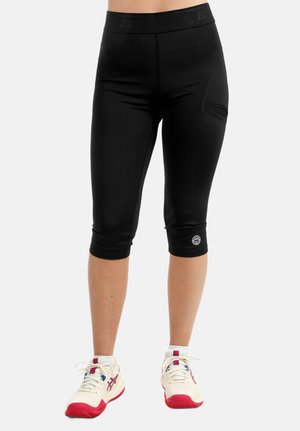 Leggings de sport noirs longueur genou avec un petit logo près de l'ourlet, portés avec des chaussures de sport blanches et rouges sur un fond neutre.