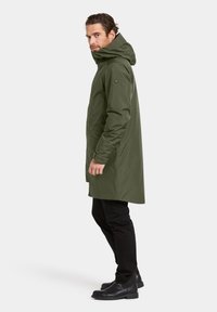 Olivengroene, knielange parka met een capuchon, voorzien van een ritssluiting en zijzakken. Draagt zwarte broek en zwarte laarzen.