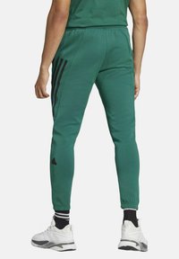Pantaloni da jogging verdi con strisce laterali nere, dotati di cintura elasticizzata e design a gamba affusolata, abbinati a sneakers bianche.