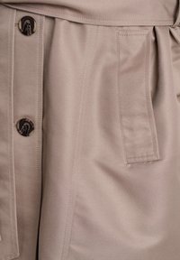 Lichtbeige stof met een gladde textuur, grote bruine knopen en een zijzak met nette stiksels en geraffineerde plooien.