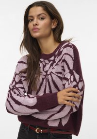 Vrouw met lang bruin haar, gekleed in een bordeauxrode en lichtroze gepatternde trui, zwarte jeans en een rode riem, poseert met gekruiste armen.