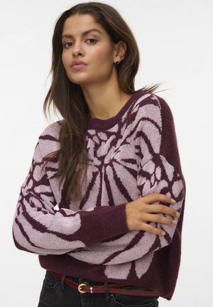 Vrouw met lang bruin haar, gekleed in een bordeauxrode en lichtroze gepatternde trui, zwarte jeans en een rode riem, poseert met gekruiste armen.