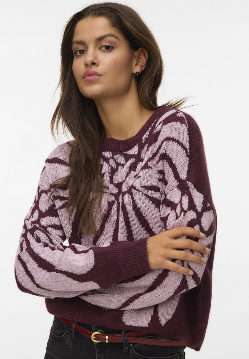 Vrouw met lang bruin haar, gekleed in een bordeauxrode en lichtroze gepatternde trui, zwarte jeans en een rode riem, poseert met gekruiste armen.