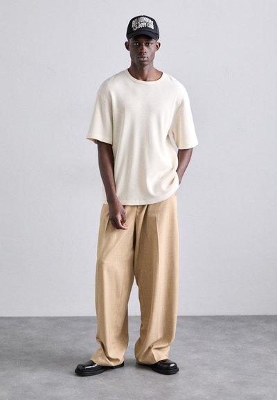 Hellbeige, übergroßes, strukturiertes Hemd, kombiniert mit lockeren, tanfarbenen Hosen und schwarzen Schuhen. Eine schwarze Kappe mit weißen Buchstaben rundet das Outfit ab.