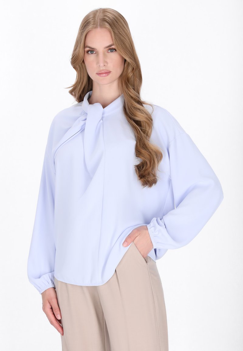 Helleblaue Bluse mit lockerer Passform, gefälteltem Detail am Kragen und elastischen Bündchen. Kombiniert mit beigen Hosen. Glatte Stoffstruktur.