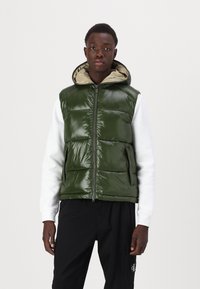 Groene pufferjas met een glanzende afwerking, ritssluiting aan de voorkant, zijzakken en een capuchon met een beige voering. Draagt over een witte langwerpige shirt met lange mouwen.