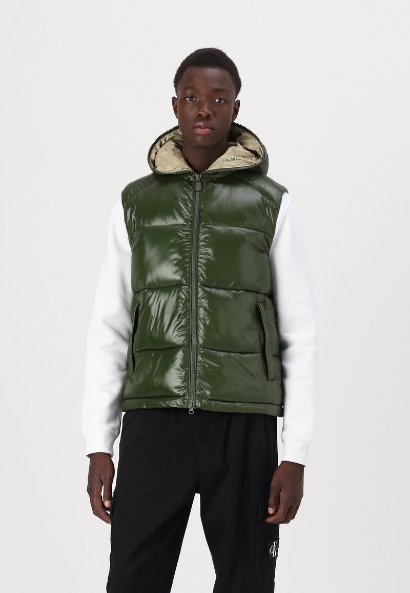 Groene pufferjas met een glanzende afwerking, ritssluiting aan de voorkant, zijzakken en een capuchon met een beige voering. Draagt over een witte langwerpige shirt met lange mouwen.