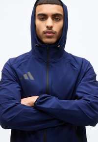 Άνδρας που φοράει μπλε σκούρο φούτερ με κουκούλα και στερεωμένο φερμουάρ, με σταυρωμένα τα χέρια, κοιτάζοντας ευθεία μπροστά πάνω σε απλό φόντο.