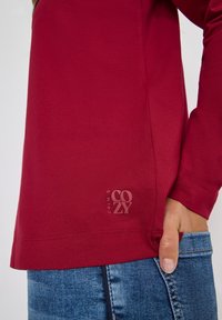 Rode longsleeve shirt van zacht materiaal, met een zichtbaar logo detail aan de zoom, gecombineerd met blauwe denim jeans.