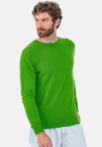 Pull vert en tissu tricoté doux avec un col rond, des manches longues, des poignets et un ourlet côtelés. Le mannequin porte un short en denim clair.