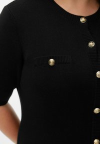 Cardigan noir en maille avec manches courtes, col en côtes, poche poitrine avec bouton doré et fermeture à boutons dorés sur le devant.