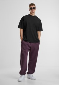 Crna oversized majica od pamuka u paru sa slobodno krojenim ljubičastim trenirkama. Model nosi bijele tenisice i sunčane naočale.