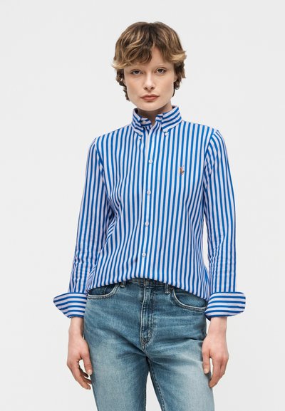 Νεαρό άτομο με κοντά μαλλιά, φορώντας μπλε και λευκό πουκάμισο με κάθετες ρίγες και ανοιχτόχρωμα μπλε τζιν, στέκεται μπροστά σε μια απλή βάση.