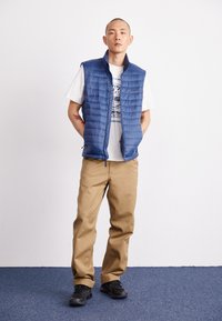 Vest albastru căptuşit, uşor, cu textură matlasată, asortat cu un tricou alb cu grafică şi pantaloni bej. Încălţăminte neagră. Poziţie în picioare.