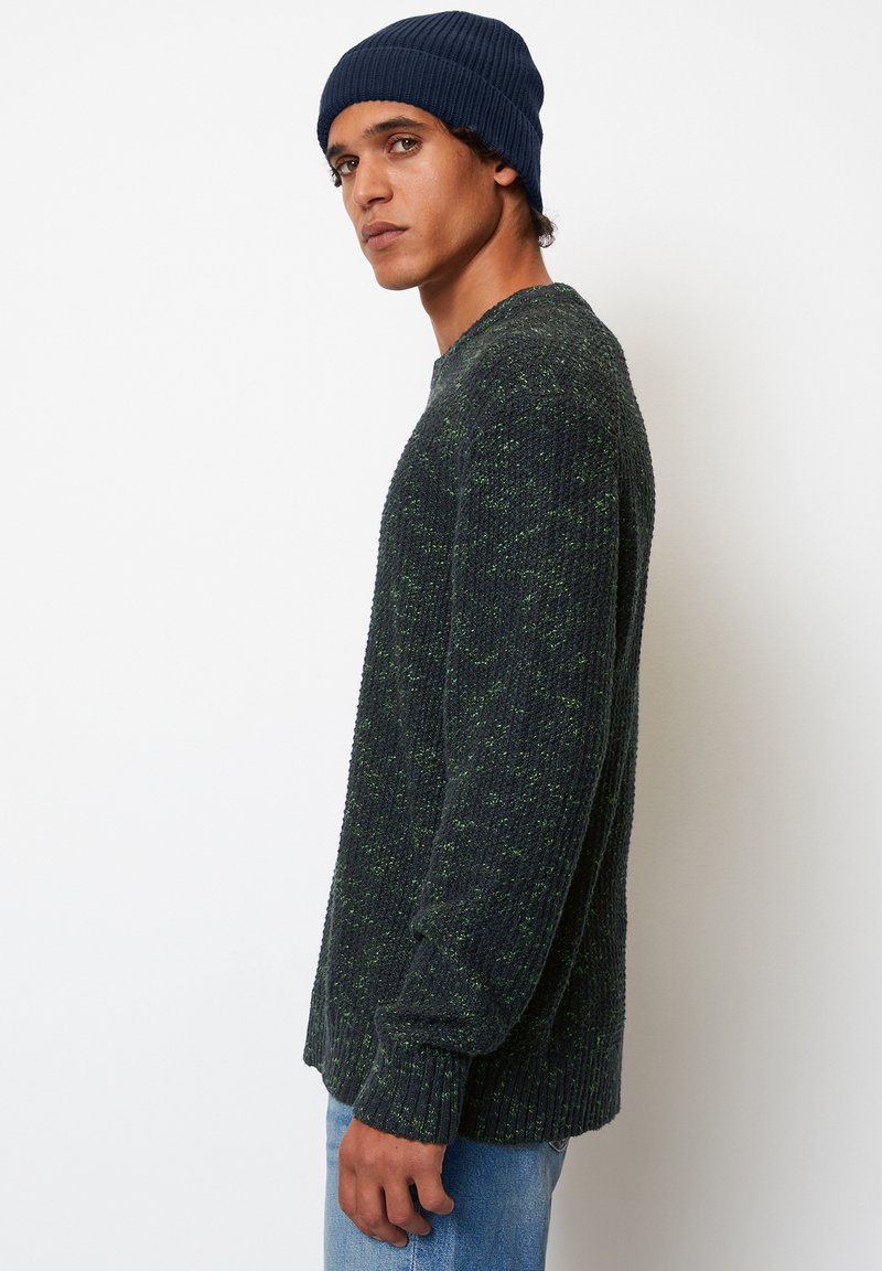 Pull en laine verte foncée avec un tricot texturé, comportant un col rond et des poignets côtelés, associé à un bonnet côtelé bleu marine et un jean bleu clair.