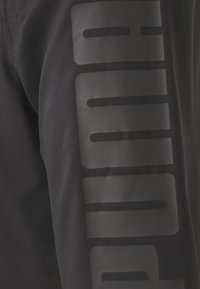 Črne športne hlače z velikim mat črnim logotipom na zunanji strani. Narejene so iz gladkega, lahkega materiala z oprijetim krojem.