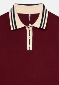 Bordeauxfärgad polo-tröja med krämfärgad krage som har två svarta ränder och en knapplist med tre knappar, även den med krämfärgad trim och svart rand.