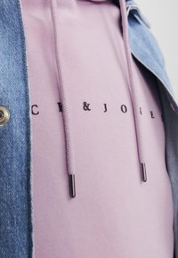 Sudadera rosa con letras negras, con capucha ajustable y puntas metálicas, llevada debajo de una chaqueta de denim con botones plateados.