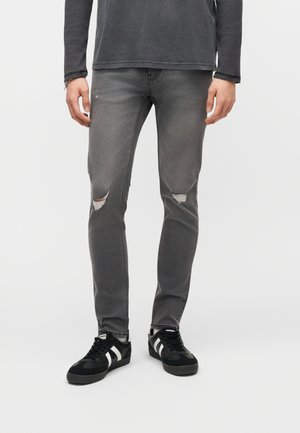 Homme portant un jean slim délavé gris déchiré et des baskets noires à rayures blanches, debout les mains détendues sur les côtés.