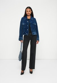 Giacca in denim a costine blu scuro con tasche frontali, abbinata a pantaloni neri a gamba larga e una borsa azzurra chiara. Tacchi neri completano il look.