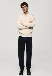 Helles beiges Zip-Knit-Pullover, schwarze gerade geschnittene Jeans und schwarze Boots. Der Stoff ist glatt, mit gerippten Bündchen und Saum.