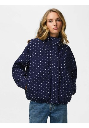 Ung kvinde med bølget blondt hår iført en mørkeblå quiltet jakke med hvide polkaprikker og blå jeans, hænderne i lommerne, neutral baggrund.