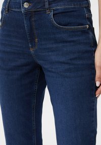 Granatowe dżinsy z pomarańczowymi przeszyciami, pięcioma kieszeniami i metalowym zapięciem. Gładka tekstura z niewielką elastycznością.