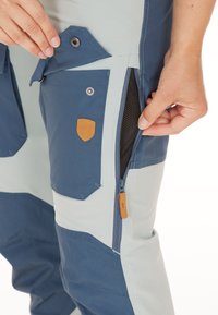 Tweekleurige werkbroeken in lichtblauw en marineblauw, voorzien van meerdere zakken, een met rits afsluitbare zijventilatie en leren details aan de voorkant.