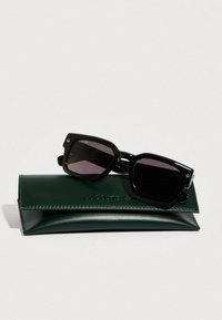 Mustat aurinkolasit, joissa on rohkea kuusikulmainen muoto, tummat linssit ja kultaiset yksityiskohdat. Ne lepäävät vihreällä nahkaisella kotelolla, johon on kohokuvioitu "LACOSTE."