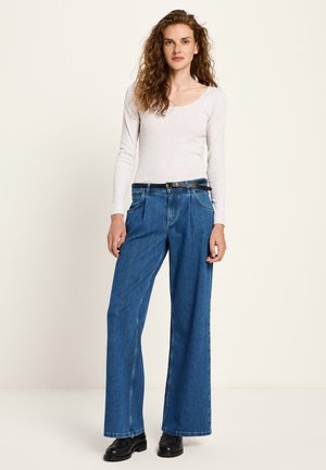 Frau mit lockigem Haar, die ein weißes Langarmoberteil, hochtaillierte, weit geschnittene blaue Jeans, einen schwarzen Gürtel und schwarze Slipper trägt und steht.