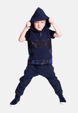 Małe dziecko stojące z rozstawionymi nogami, ubrane w granatową bezrękawnikową bluzę z wzorem galaktyki, granatowe spodnie i granatowe wsuwane buty, zakładające kaptur na głowę.