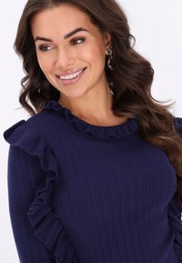Granatowy sweter prążkowany z falbankami na ramionach. Posiada szeroki dekolt i strukturalny materiał. Modelka prezentuje uśmiech.