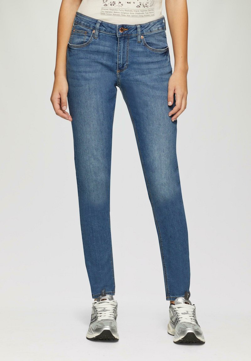 Schmale, mittelhohe blaue Jeans mit leichter Ausbleichung; aus Denim gefertigt, mit Fronttaschen und Akzenten aus Kupferhardware; kombiniert mit silbernen Sneakers.