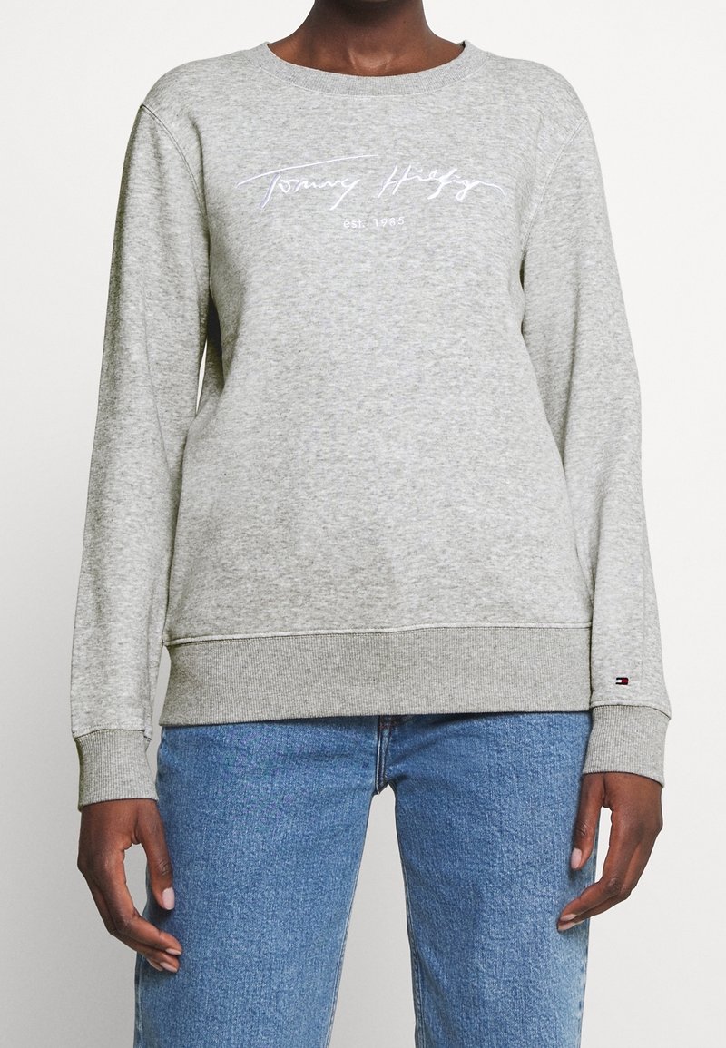 Personne portant un sweat-shirt gris Tommy Hilfiger avec le logo sur la poitrine et un jean bleu, debout devant un fond uni.